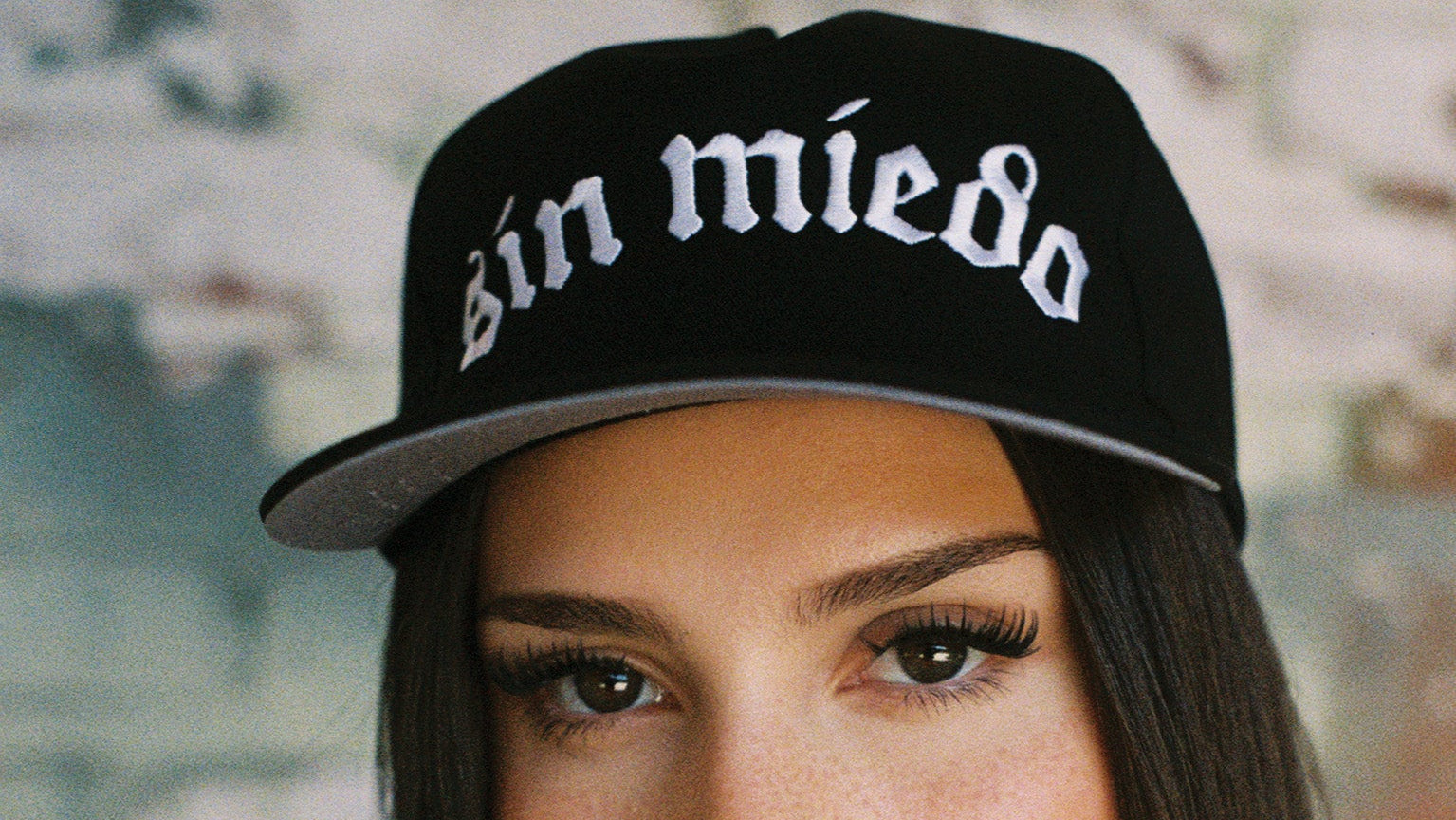 A woman wearing a black baseball cap with the Spanish phrase 'sin miedo' in white Gothic-style lettering, posed against a blurred brick wall background.